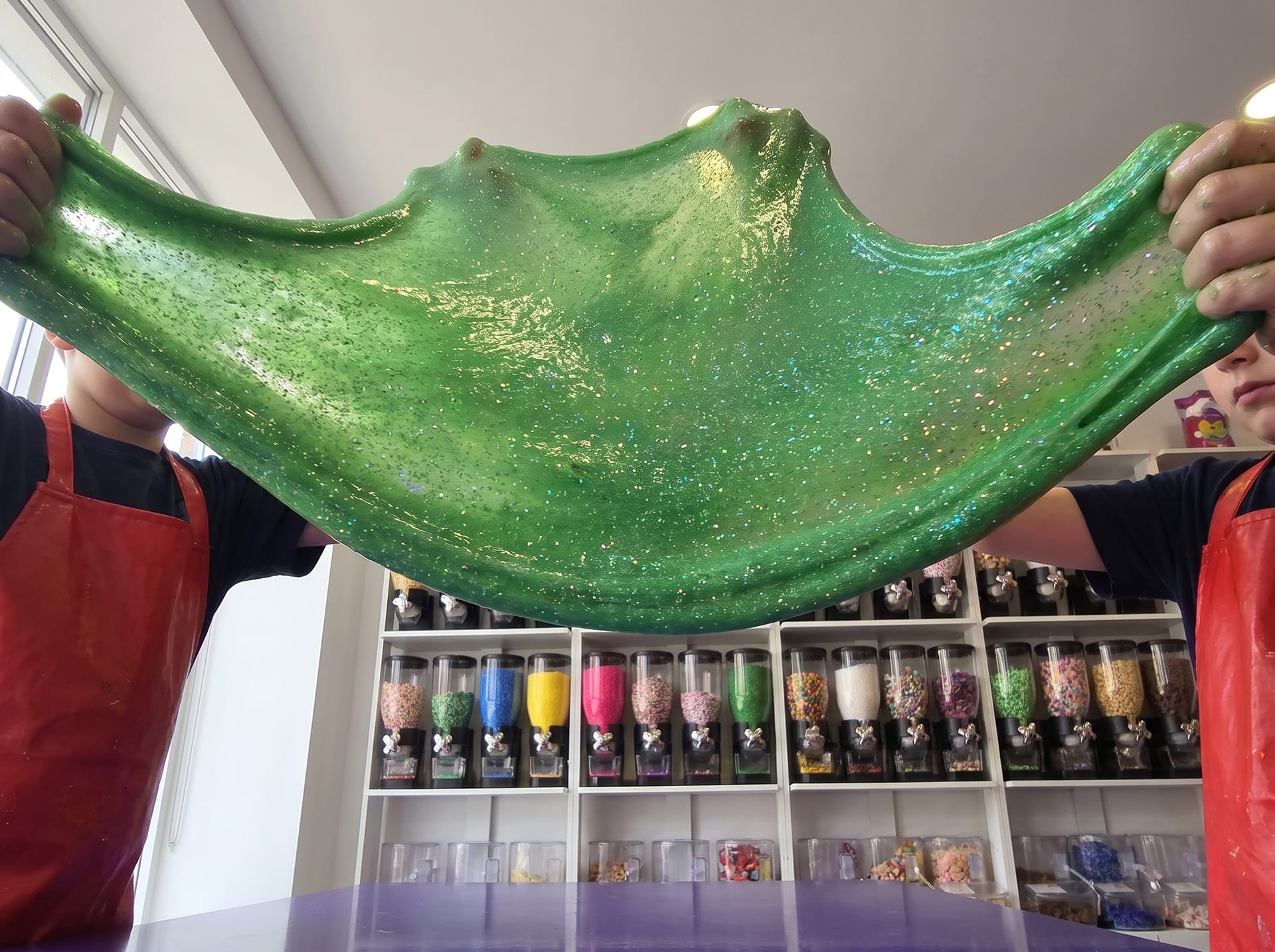 A wide sheet of glittery green slime stretched between two pairs of hands
